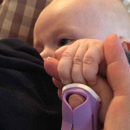 Baby’s hand gripping a caregiver’s finger while a purple Thumble files nails, by Thumble Baby Care.