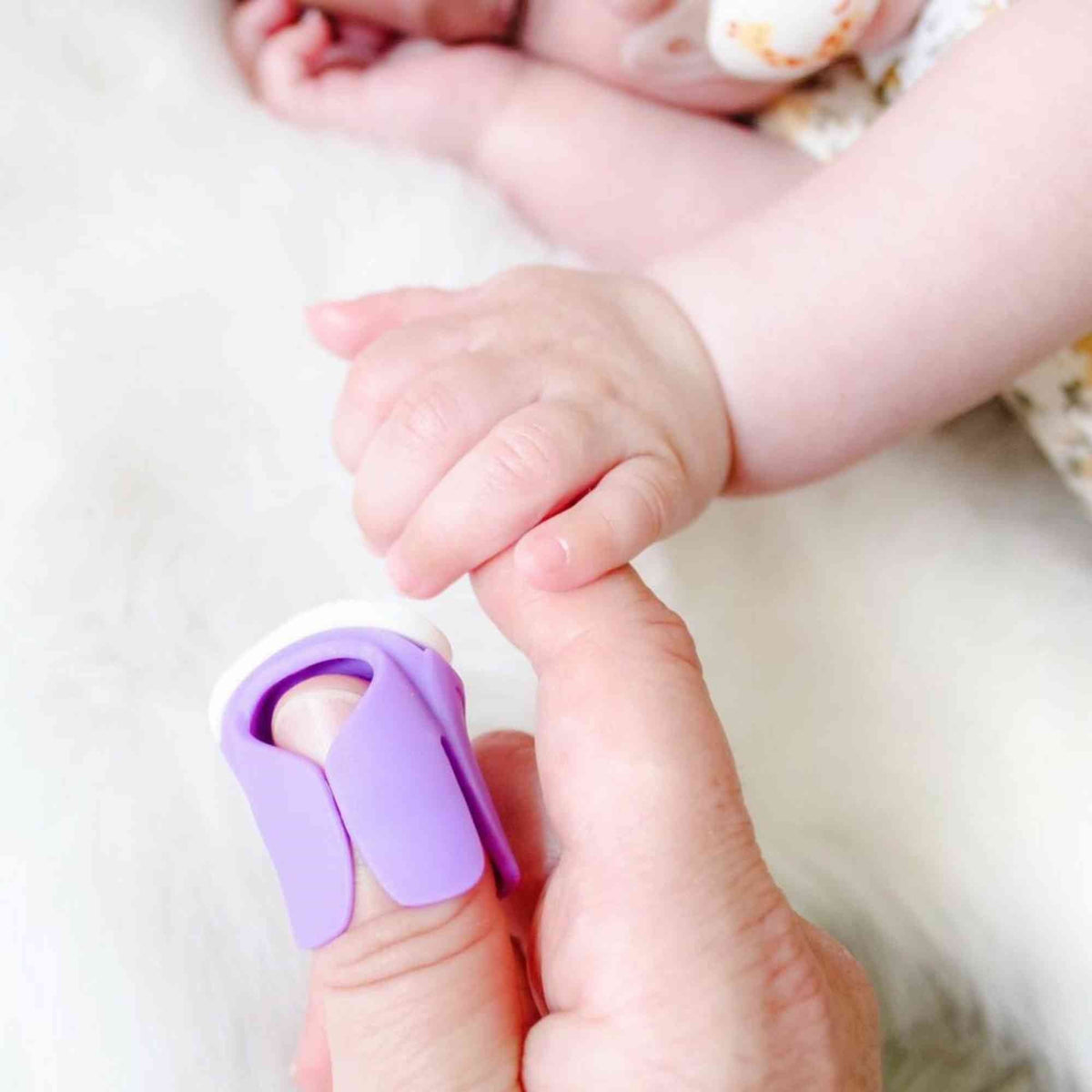 Purple Thumble on an adult thumb beside a sleeping baby’s hand, by Thumble Baby Care.